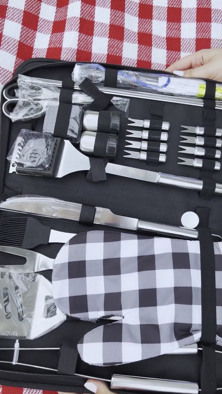 Tampilan close-up dari atas tangan yang membuka satu set alat barbekyu portabel hitam di atas selimut piknik kotak-kotak merah. Kit ini termasuk aksesori panggangan baja tahan karat dan sarung tangan oven untuk memasak di luar ruangan.
