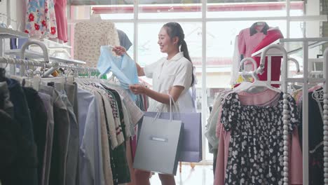 young asian woman with the shopping bags choosing blue clothing item then smiling to the camera while shopping at the boutique