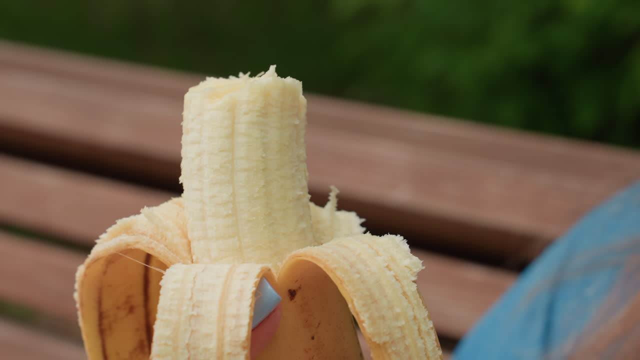 여자가 공원 벤치에 앉아 싱싱한 녹음으로 둘러싸여 날것의 플랜테인을 먹는 모습의 클로즈업 사진이에요. 부드러운 자연광이 편안한 분위기와 과일의 질감을 강조하고, 평온한 야외의 순간을 담아냈어요.