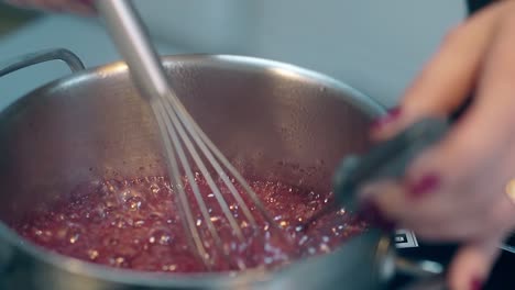 woman stirs red boiling caramel in saucepan using whisk