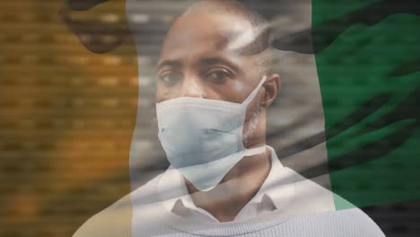 animation of flag of ivory coast waving over african american man wearing face mask in city street