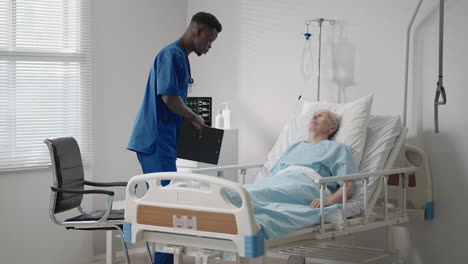 an elderly male patient lying in a hospital bed connected to an ecg machine talks to a black doctor in a protective mask