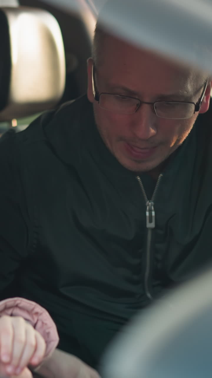 un hombre con una chaqueta verde está desabrochando cuidadosamente el cinturón de seguridad de su hija joven, que está vestida con una jaqueta rosa y una gorra rosa a juego, dentro de un coche