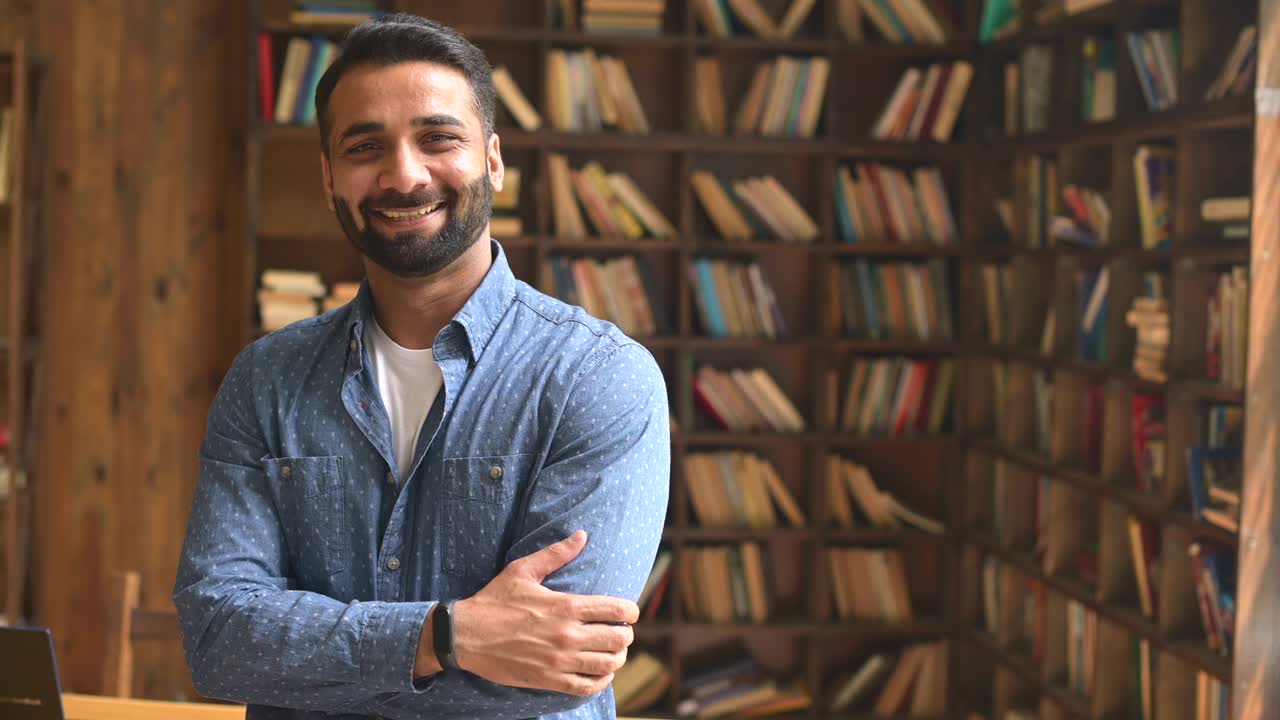 un empresario indio barbudo sonriente se para cerca del escritorio, mira hacia otro lado y luego a la cámara.