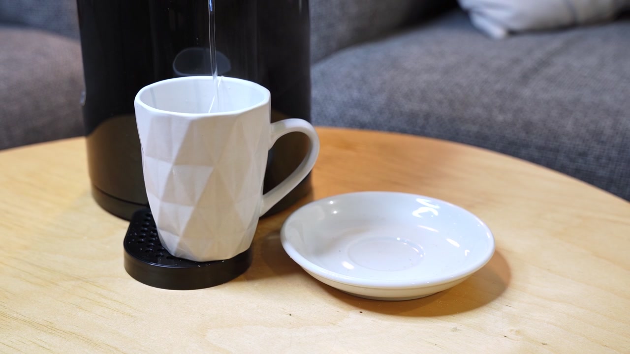 Nærbilde av en kvinnes hender som plasserer et hvitt geometrisk krus under en svart kaffemaskin på et trebord. Klart varmt vann helles i den keramiske koppen i et moderne stuemiljø