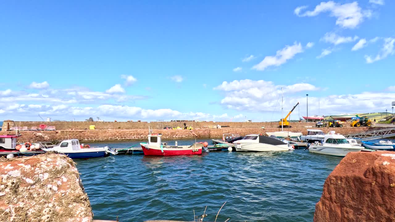 Several small boats gently float near a stone pier in a coastal town harbor under bright daylight, with calm water and clear blue skies