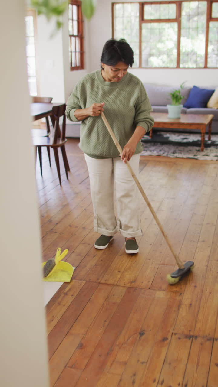 video vertical de una mujer biracial mayor limpiando el suelo en la sala de estar