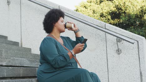 mujer biracial feliz de talla plus usando un teléfono inteligente y bebiendo café