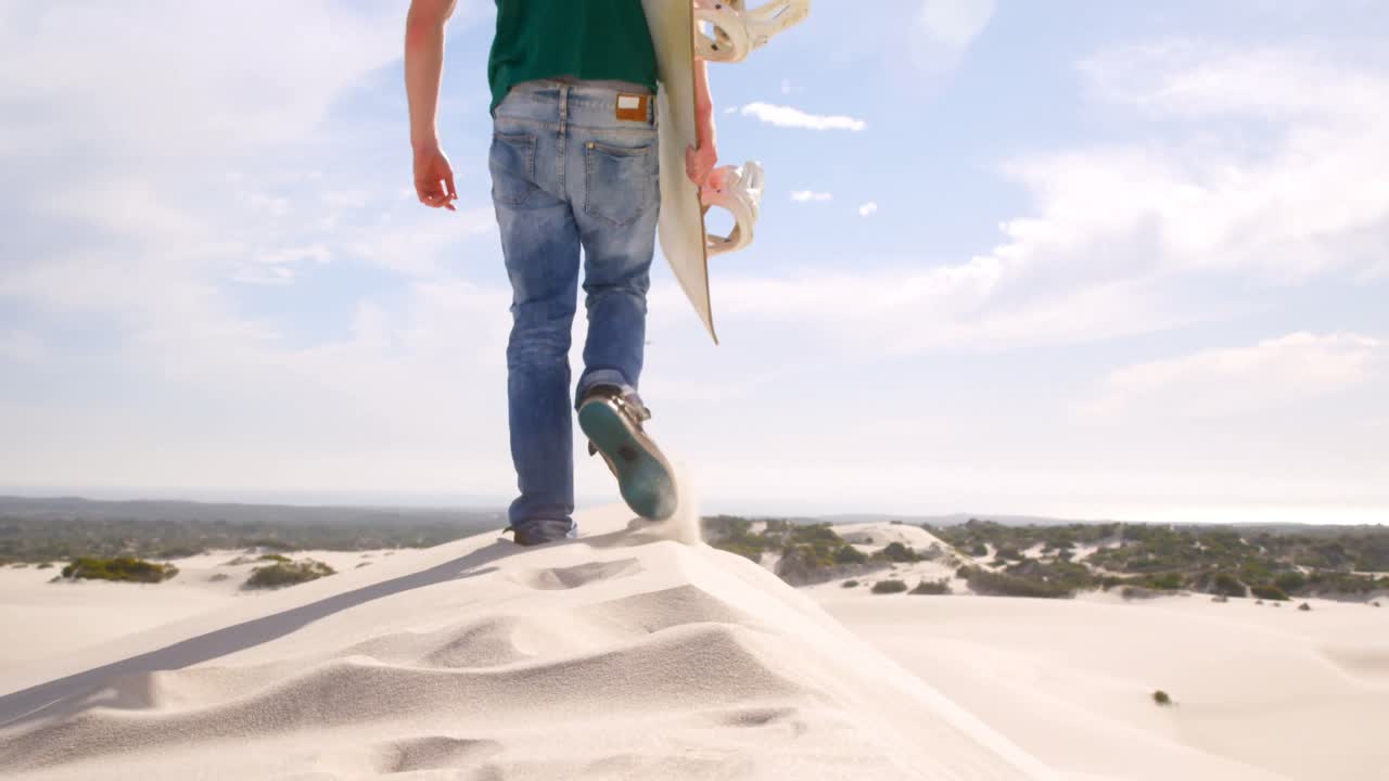 Uomo che cammina con tavola da sandboarding nel deserto 4k