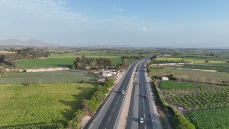 Aerial-drone-shot-of-vehicles-crossing-four-lane-Panamericana-Norte-highway-surrounded-by-mountains-in-Peru-through-farming-fields-on-both-corners-on-a-sunny-day