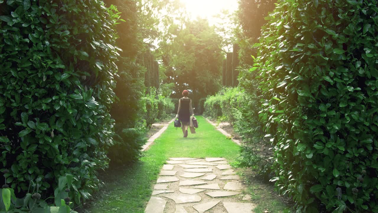 A person walks down a sunlit garden path between lush green hedges, carrying bags.