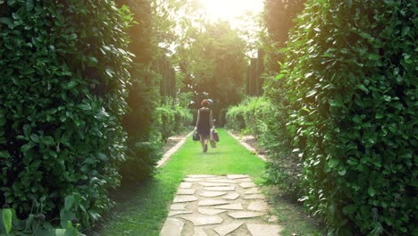 a person walks down a sunlit garden path between lush green hedges, carrying bags.