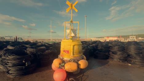 una robusta boya de navegación amarilla se alza en el muelle de un pueblo pesquero costero español, actuando como un faro de seguridad marítima y orientación en medio de las animadas actividades portuarias.