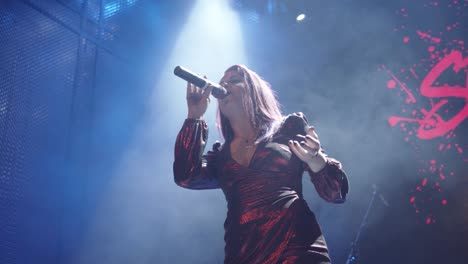 a female performer wearing a red dress singing into a cordless microphone against a blue stage with a spotlight shining from above in a smoke-filled room