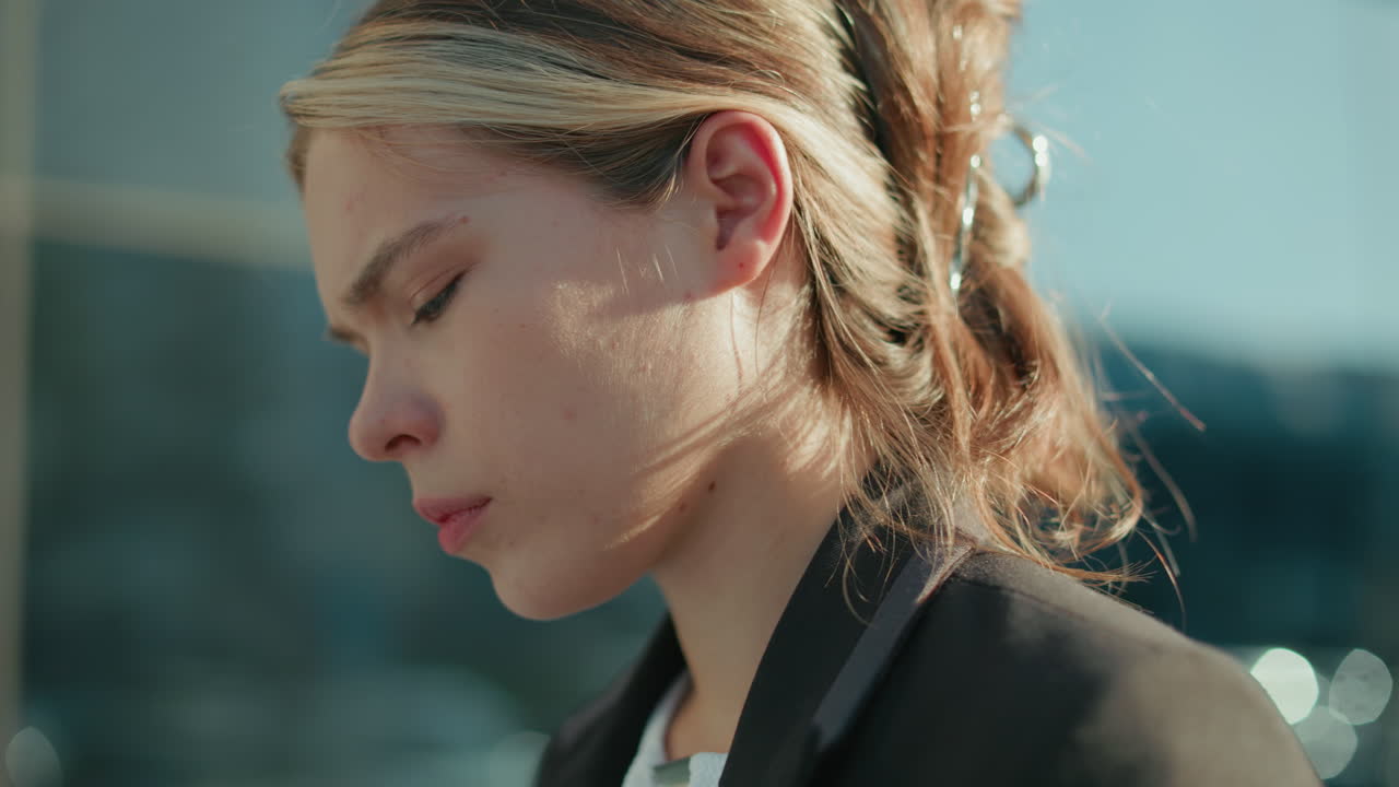 Young professional woman with serious expression focused intently on task outdoors near glass building under sunlight, showcasing concentration and determination in urban business environment