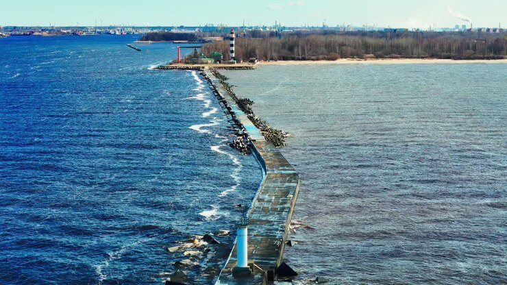 A narrow concrete pier with a lighthouse at the end stretches into the Baltic Sea, guiding vessels toward the coast, with an industrial cityscape in the background
