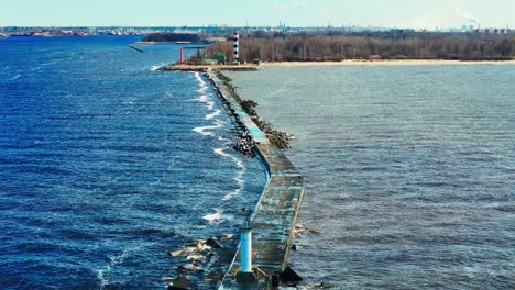 a narrow concrete pier with a lighthouse at the end stretches into the baltic sea, guiding vessels toward the coast, with an industrial cityscape in the background