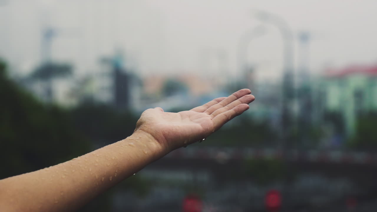 Close-up of a girl's hand reaching out to catch falling raindrops against a blurry city background. Moody, atmospheric footage of rainy urban weather and sensory connection with nature