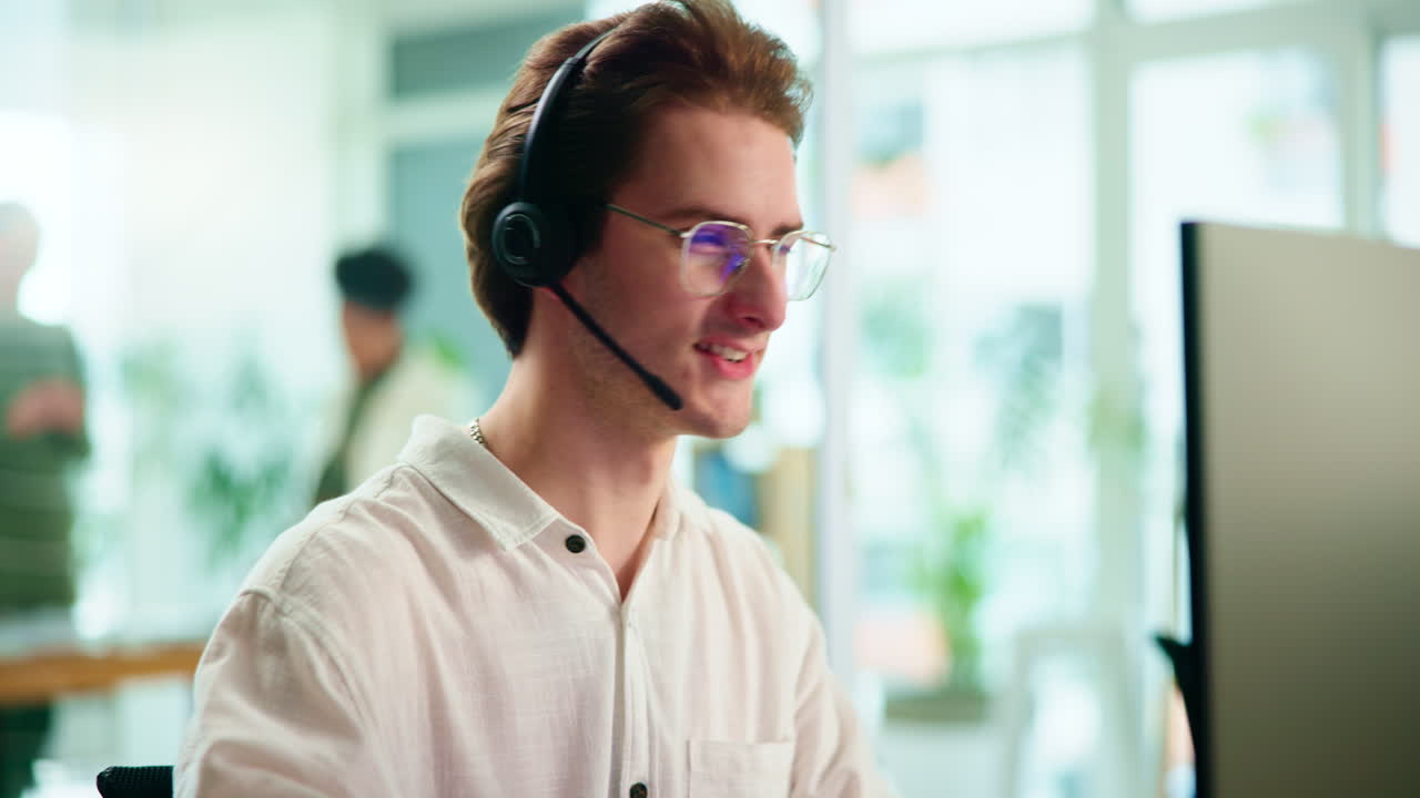 Young man working in a call center, wearing a headset and smiling while talking to a customer
