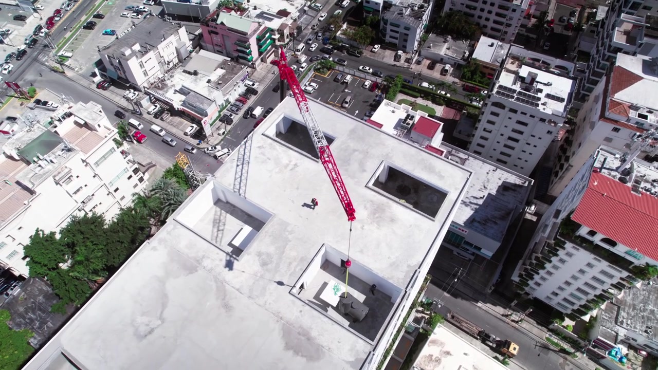 Toma aérea de dron en picado orbitando a la izquierda sobre un edificio en construcción mientras una grúa móvil roja baja un tanque de agua en una abertura en el tejado en Santo Domingo.