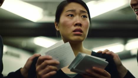 a young woman stands in a shadowy area, her expression filled with concern as she clutches several papers. two individuals approach her, trying to console or communicate something important.