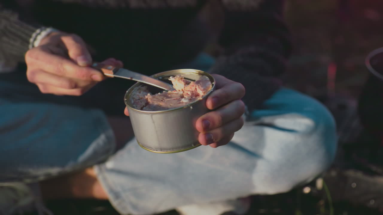 close up of person eating canned food with knife seated outdoors enjoying meal in natural forest setting casual clothing camping activity picnic leisure fresh green grass daylight happiness youth