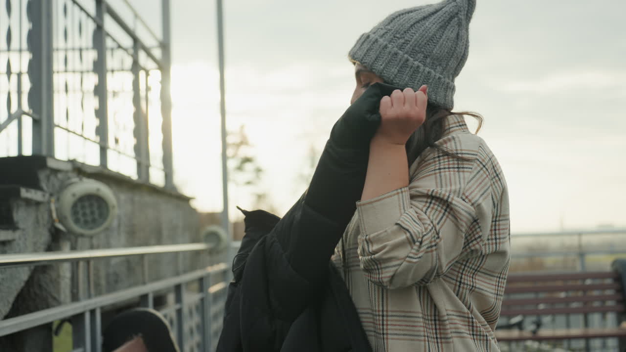 Woman removing jacket on rooftop stairs at sunset, playful freestyle dance and groove, adjusting beanie, loose rhythm, expressive hand gestures, casual plaid outfit, warm backlight, candid performance