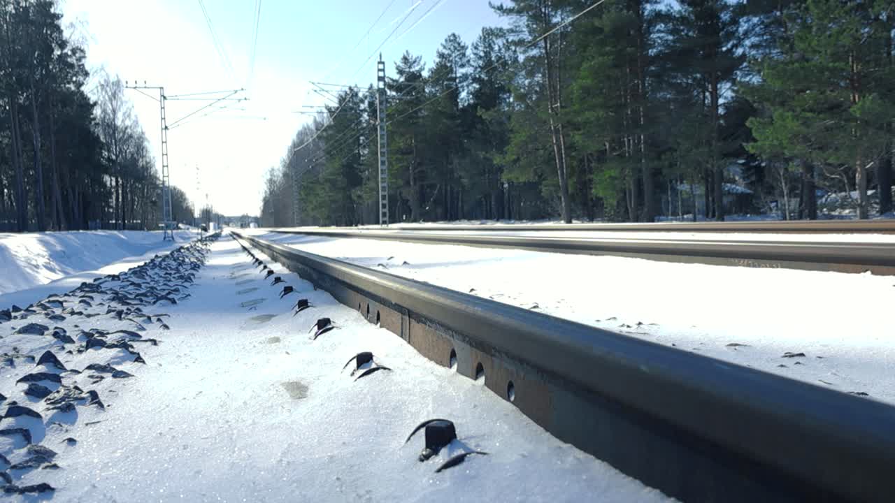 Low angle panning of electric trains railroad tracks covered in snow stretching into the distance under the winter sun.
Scenic electric multiple unit rail roadside bordered by stoic evergreen trees