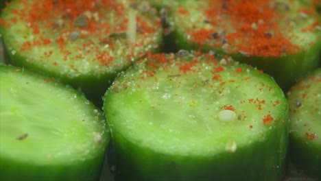 a macro close up shot, falling sesame seeds grains on a cut slices of a fresh salted green cucumber with red sweet paprika on it, slow motion 4k video