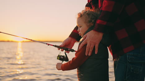 Feliz-Niño-Rizado-Está-Pescando-Con-Caña-De-Pescar,-El-Padre-O-El-Abuelo-Están-Ayudando-A-Enseñar-A-Su-Hijo-A-Pescar