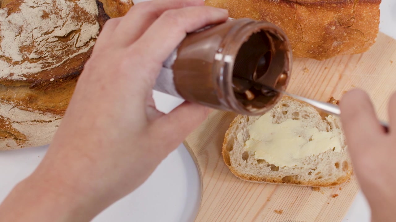 Primo piano di mani che usano un coltello per spalmare crema al cioccolato sui toast della colazione.