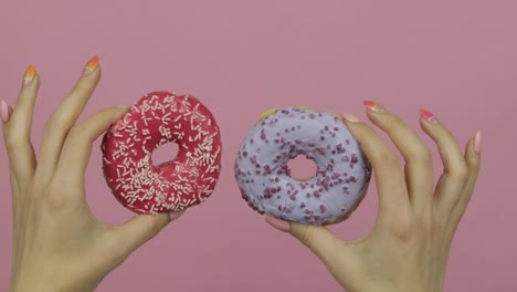 womens hands holding two red and blue, sprinkled donuts on pink background.