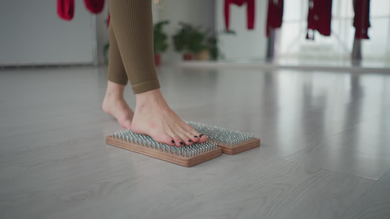 Young wellness guide balances barefoot foot on wooden nail mat in serene yoga studio with red aerial silks and bright windows practicing acupressure therapy for relaxation and inner calm grounding