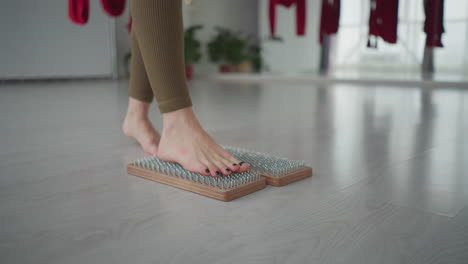 young wellness guide balances barefoot foot on wooden nail mat in serene yoga studio with red aerial silks and bright windows practicing acupressure therapy for relaxation and inner calm grounding