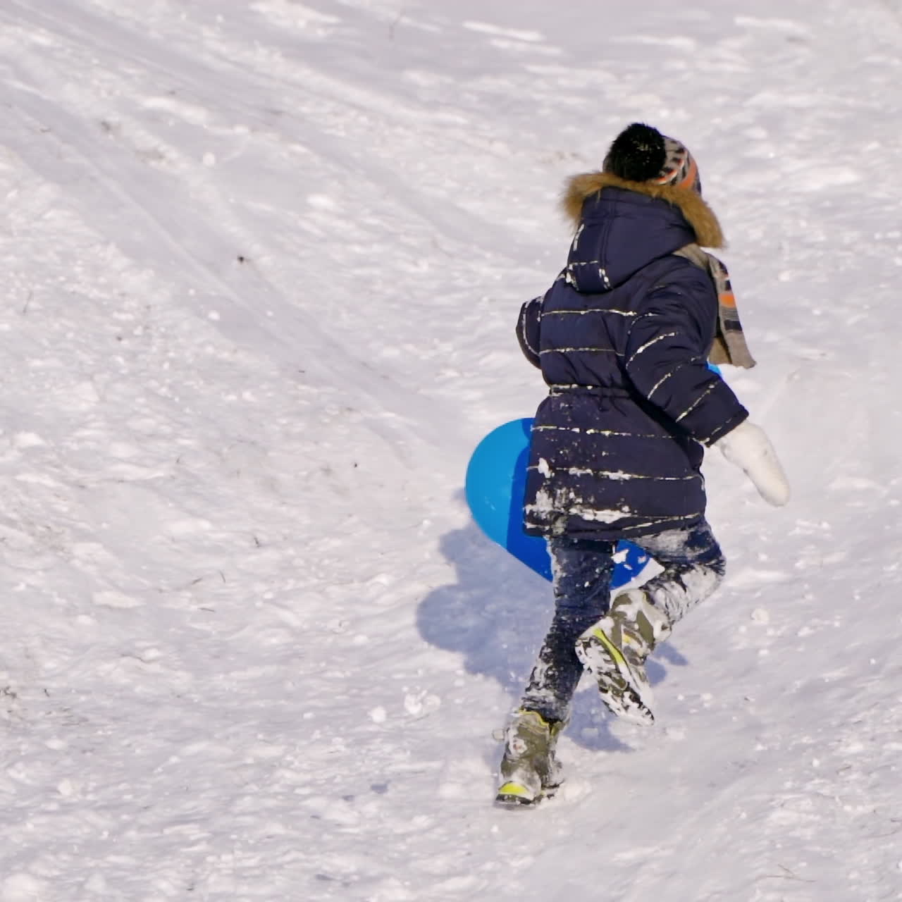 Children on snow in winter. Little boy rising up the hill with his plastic plate outdoors. Joyful childhood during winter vacation. Slow motion.