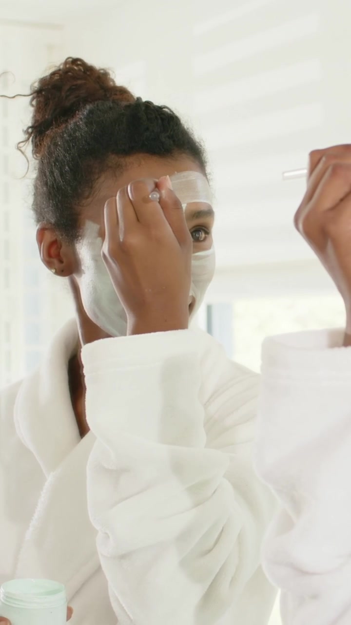 Vertical video: Loading brush, applying mask, woman in robe at vanity using gold brush to prep skin