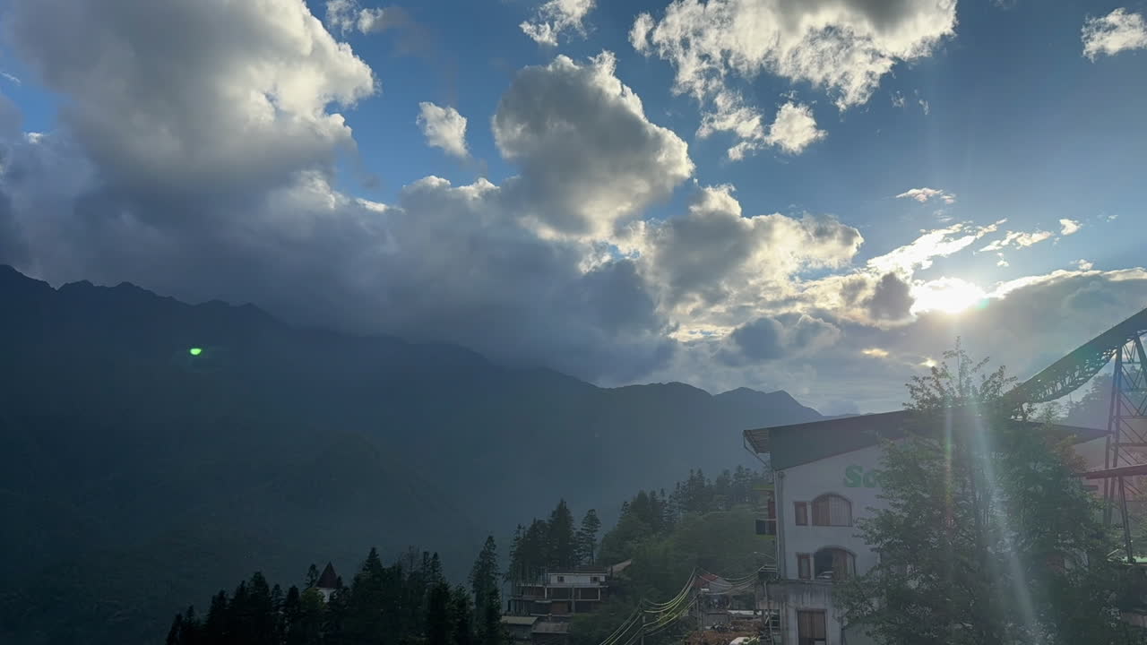 Sunlight rays pierce through fast-moving clouds above a misty mountain landscape. Fixed shot captures the dramatic light shift.