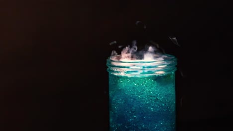 a close-up of swirling smoke rising from a jar filled with shimmering blue liquid against a dark background.