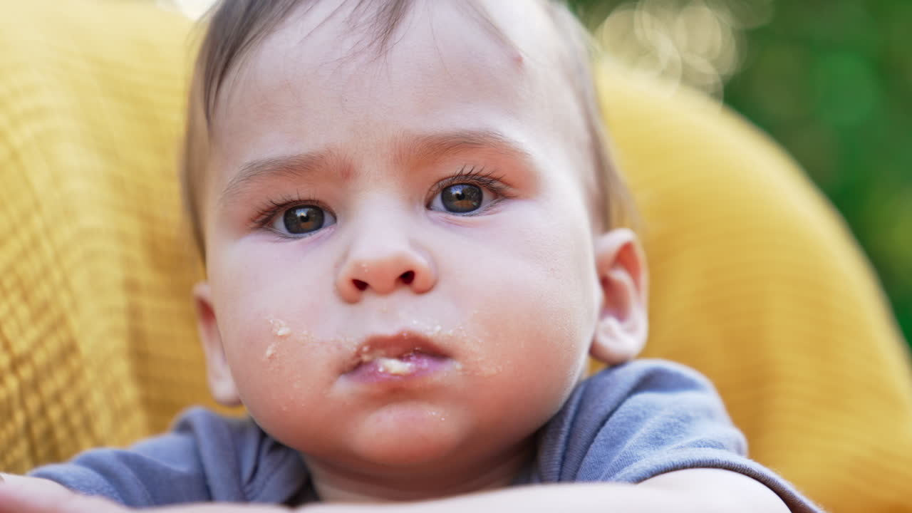 Serious cute toddler with smudged face looks calmly into camera. Mom gives full spoon to a son but he turns the head away. Close up.