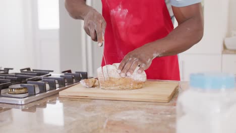 sección media de hombre afroamericano con delantal y cortando pan en la cocina