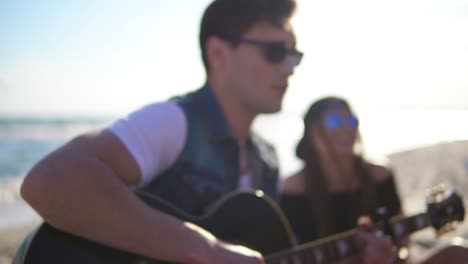 joven tocando la guitarra entre un grupo de amigos sentados en sillas fáciles en la playa y cantando en una noche de verano. disparo en cámara lenta