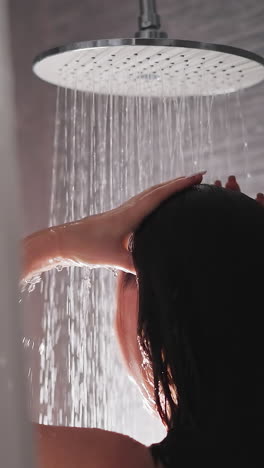 mujer morena lava el cabello y el cuerpo en la ducha. dama acaricia el cabello en la ducha moderna con azulejos grises. agua de la ducha grande fluye por el cuerpo de la dama