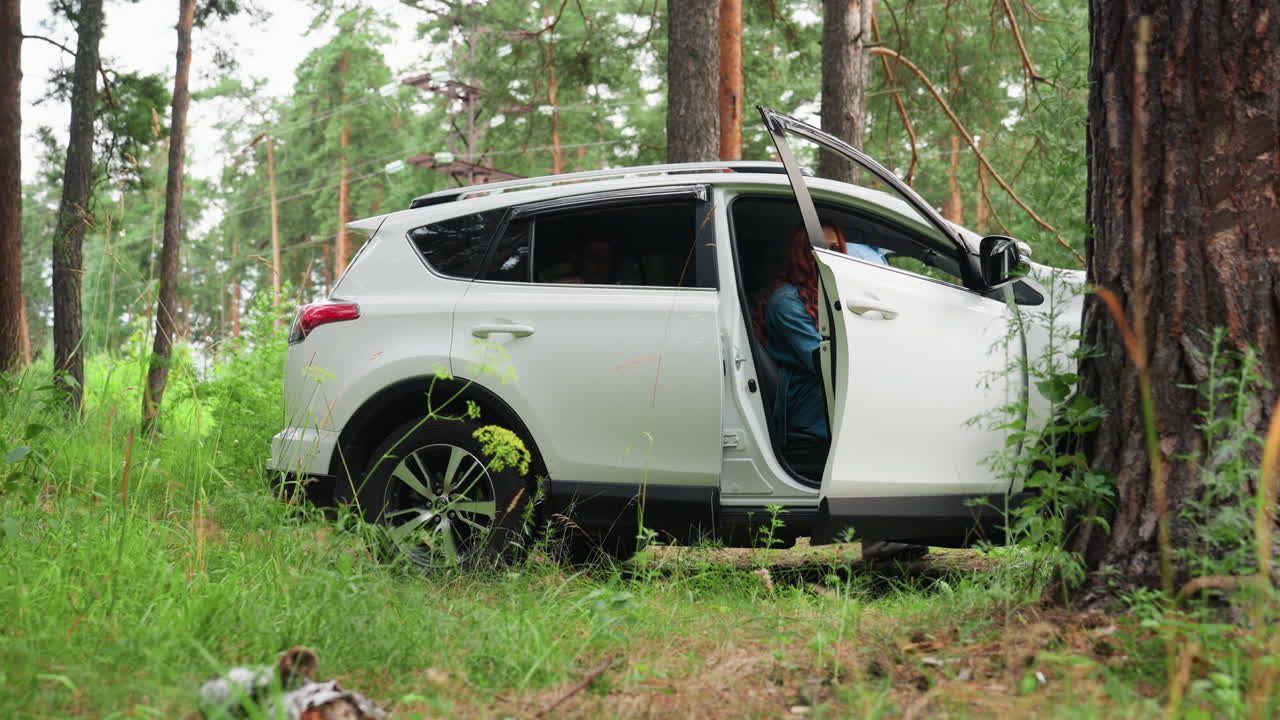 Family gets into white suv for vacation in forest clearing, mother climbs into front seat while youngest sticks head out window smiling, siblings settle inside, doors open, trees, summer departure