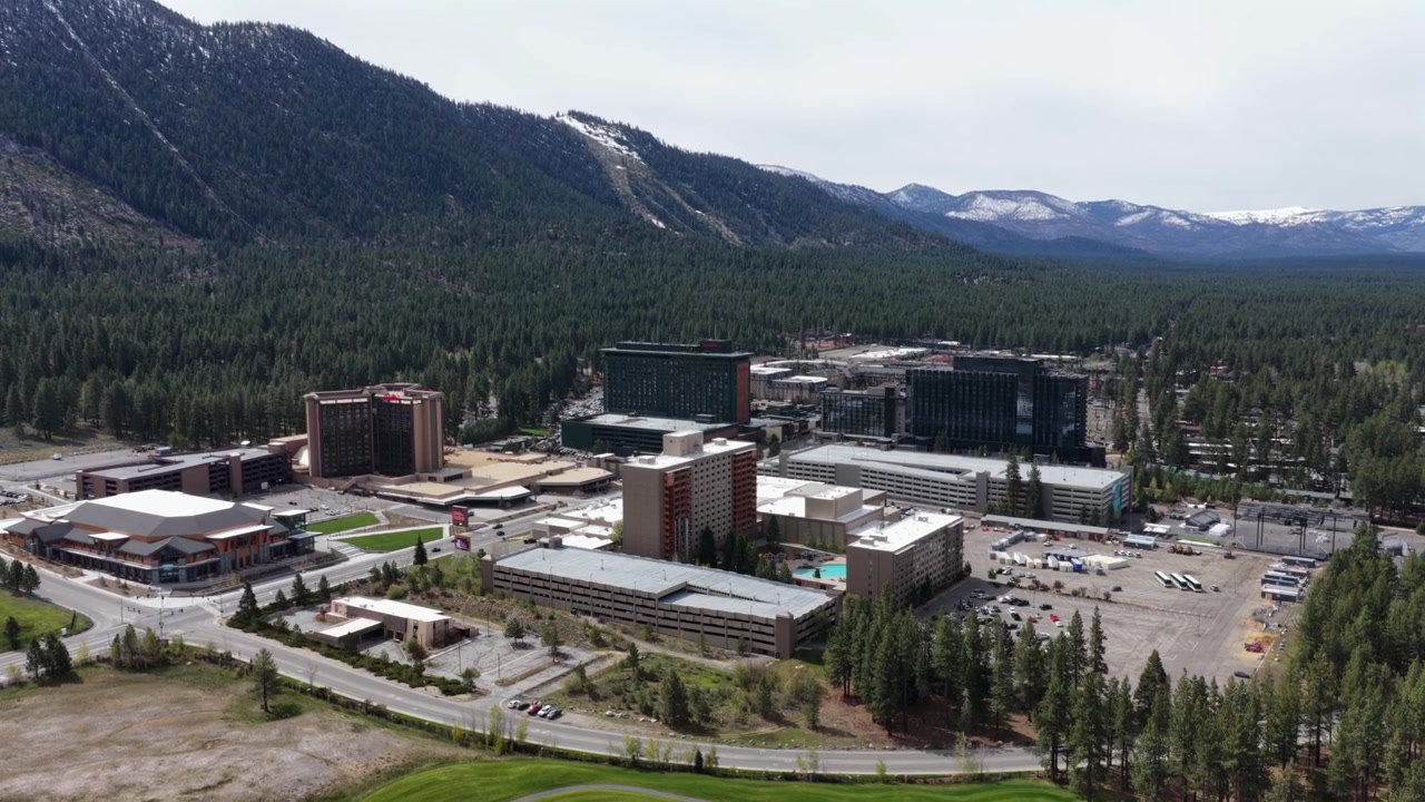 Complesso turistico con casinò e torri alberghiere a più piani accanto a un campo da golf e un'autostrada tortuosa, fitta pineta e cime della Sierra Nevada spolverate di neve che si ergono ripide dietro. Nevada, USA. Veduta aerea