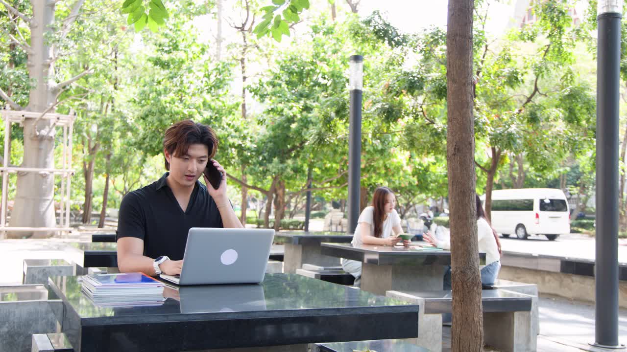 アジア系の男子学生が、大学キャンパスの屋外テーブルでノートパソコンを使い、電話で話している。