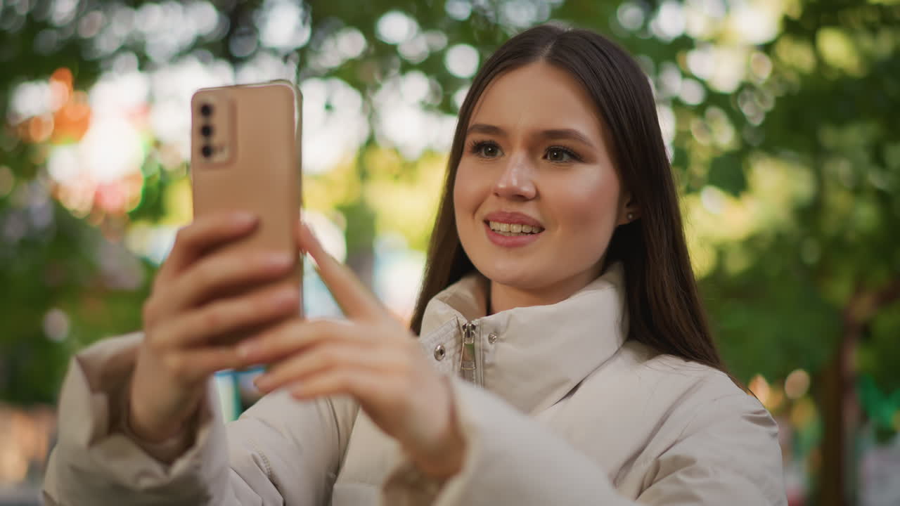 Uma jovem ao ar livre tirando uma selfie, capturando um retrato sorrindo com um smartphone em um parque arborizado cheio de bokeh e luz solar quente, usando uma jaqueta puffer leve, inclinando suavemente a cabeça, tocando na tela e gravando