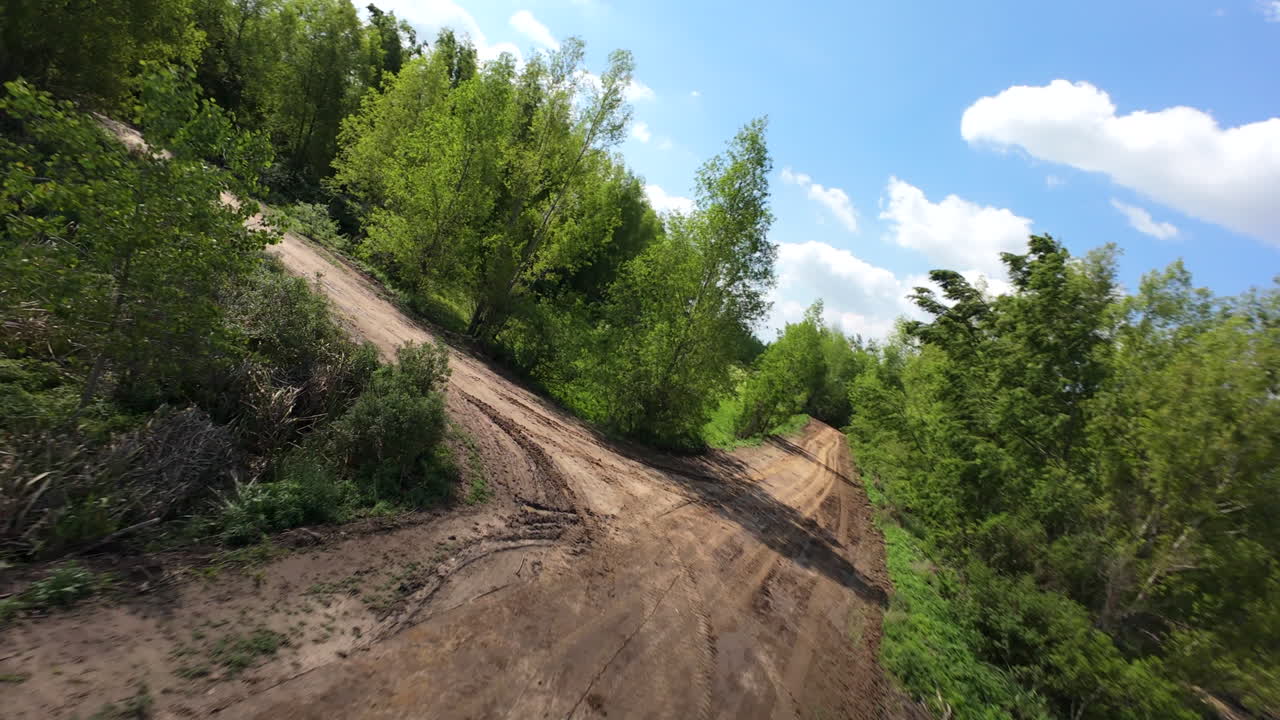 FPV drone flight low over dirt road and turning through trees on sunny day