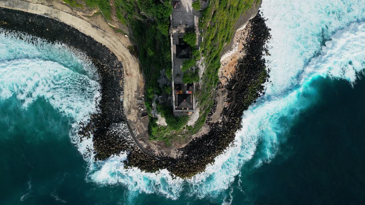 Pohled shora dronem na Uluwatu ukazující chrám poblíž okraje útesu, jasné oceánské vzory, skalnaté pobřeží a silné pobřežní vlny, ideální pro cestování, přírodu a záběry krajiny Bali