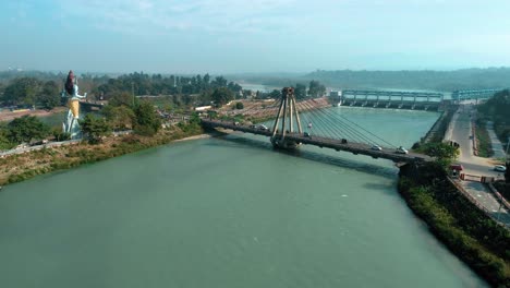 Wide-aerial-shot-of-Holy-city-Haridwar-showing-the-big-statue-of-Hindu-God-Lord-Shiva,-bridge-over-holy-Ganges-River-and-traffic-passing-by-the-bridge