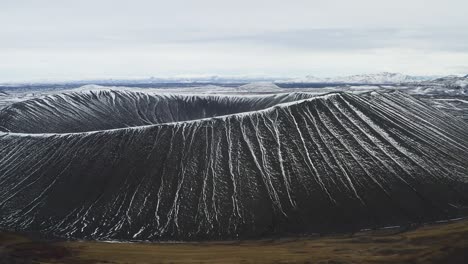 Aerial establishing shot of Hverfjall volcano crater in Iceland during cloudy morning. Snow covered turf ring. Lateral shot.